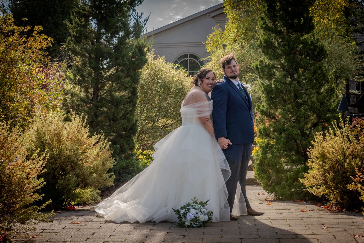 A bride and groom stand close on a stone path, surrounded by fall leaves and evergreens, bathed in warm sunlight.