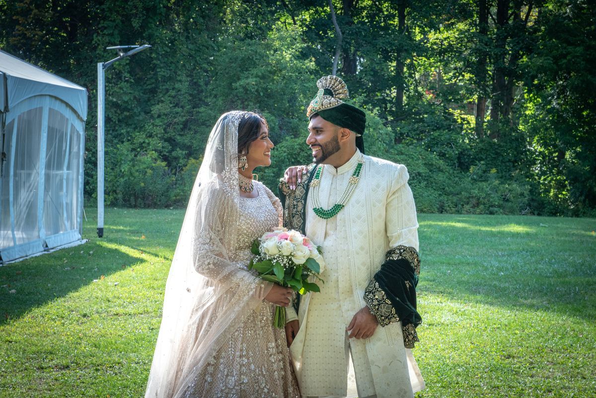 A bride and groom exchange smiles on a sunlit lawn, surrounded by trees, during a traditional South Asian wedding ceremony.