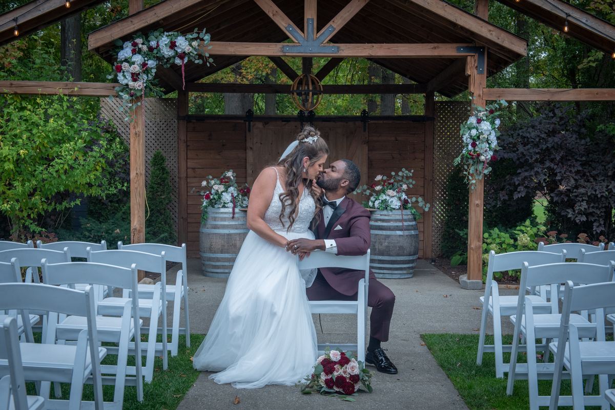 A bride and groom kiss under a wooden arbor decorated with flowers, flanked by wine barrels at an outdoor wedding.