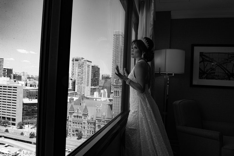 A bride in a sparkling gown and tiara stands by a window, gazing at an urban skyline, illuminated by soft natural light.