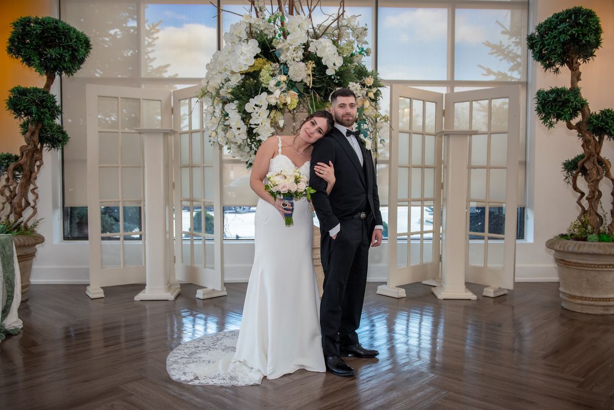 A bride and groom pose under a floral arch in a lit indoor space with topiary trees and open windows.