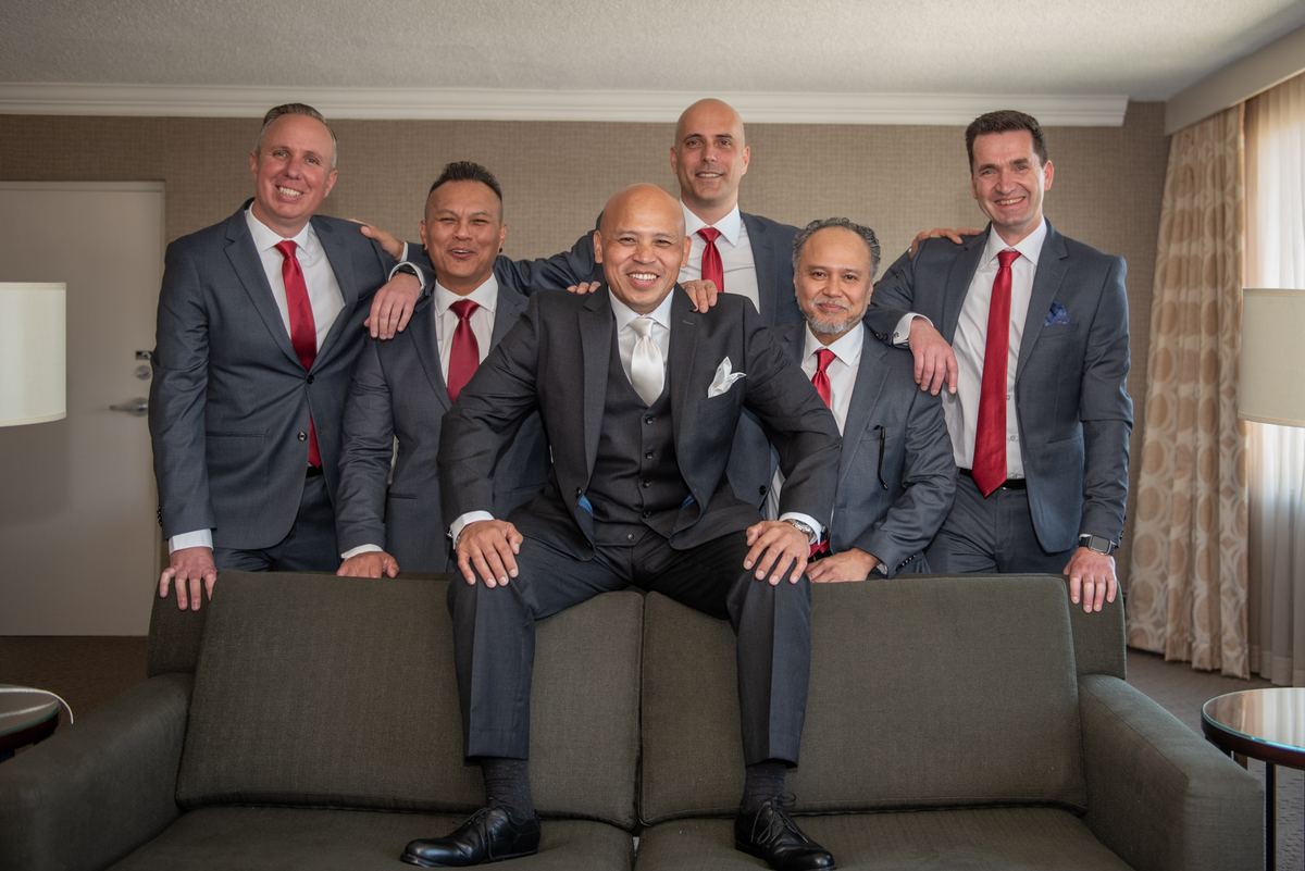 A groom and his groomsmen in matching dark suits with red ties smile warmly at the camera in a well-lit hotel room.