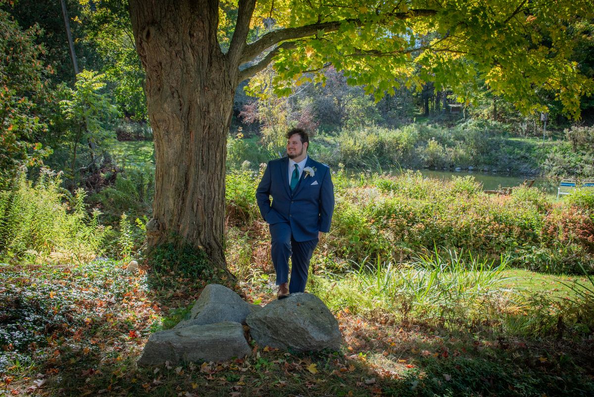 A groom in a navy suit smiles as he steps onto a rock near a large tree, surrounded by autumn leaves and a serene creek.