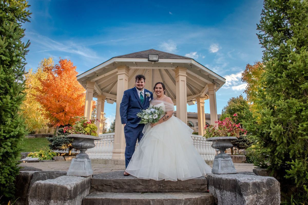 A smiling bride and groom stand on stone steps before a gazebo surrounded by autumn leaves under a clear blue sky.