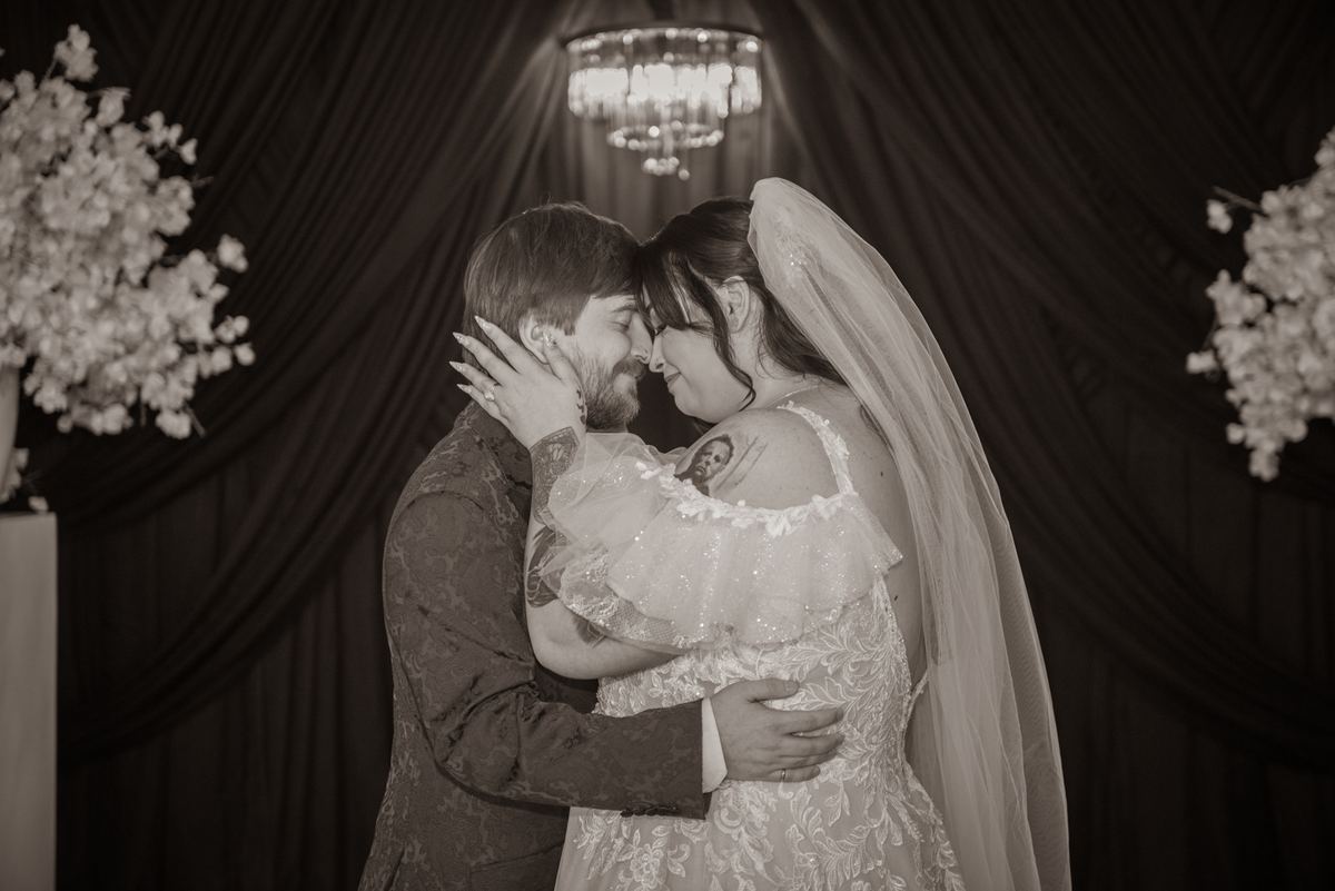 A bride and groom embrace tenderly with foreheads touching in soft light, surrounded by curtains and flowers.