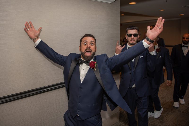 A groom in a navy tuxedo with a red boutonnière joyfully raises his arms as he walks down a corridor.