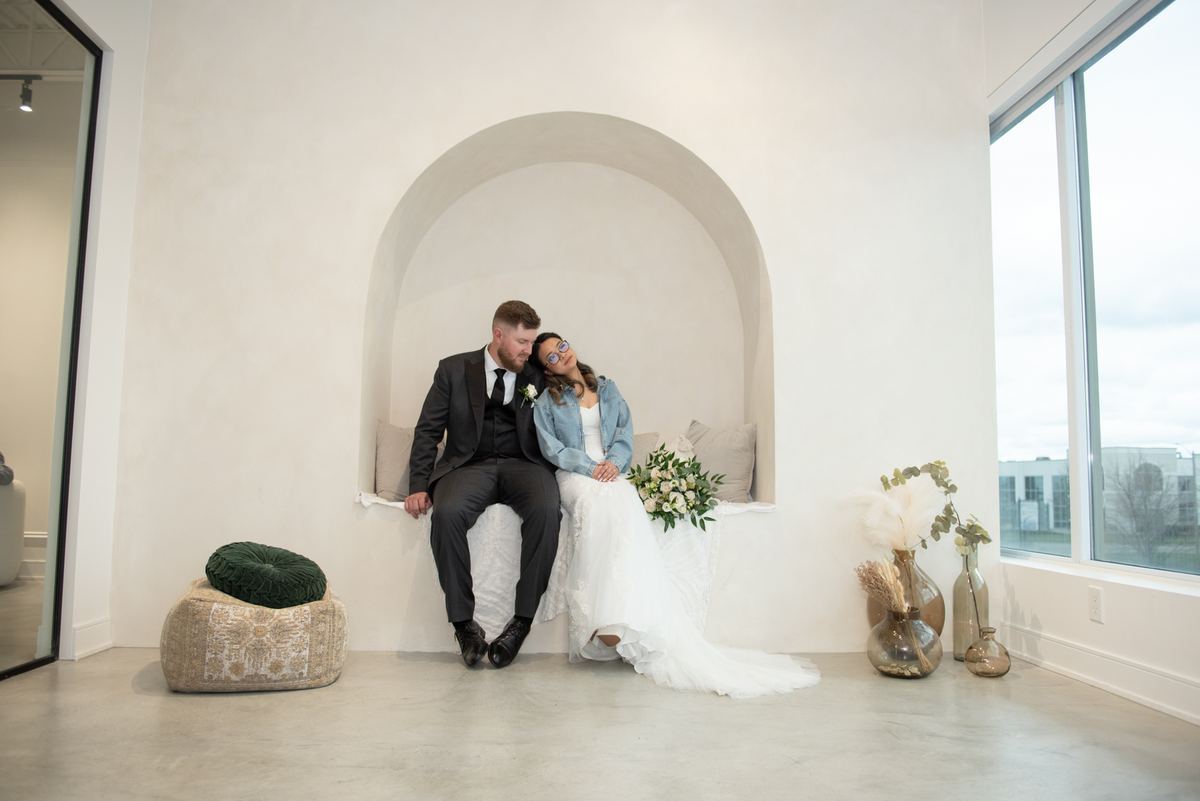 A bride in a white gown and denim jacket leans against her groom in a dark suit within an arched alcove.