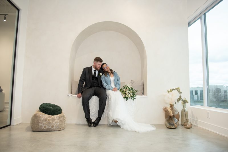 A bride in a white gown and denim jacket leans against her groom in a dark suit within an arched alcove.