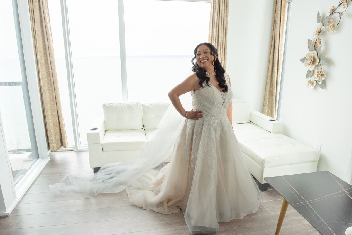 A smiling bride in a lace gown poses confidently in a modern, sunlit living room with floor-to-ceiling windows.