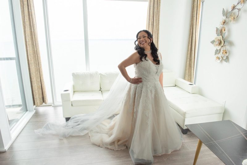 A smiling bride in a lace gown poses confidently in a modern, sunlit living room with floor-to-ceiling windows.