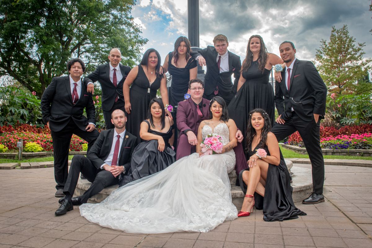 Bride in white dress with pink bouquet stands with wedding party on stone bench in garden under partly cloudy sky.