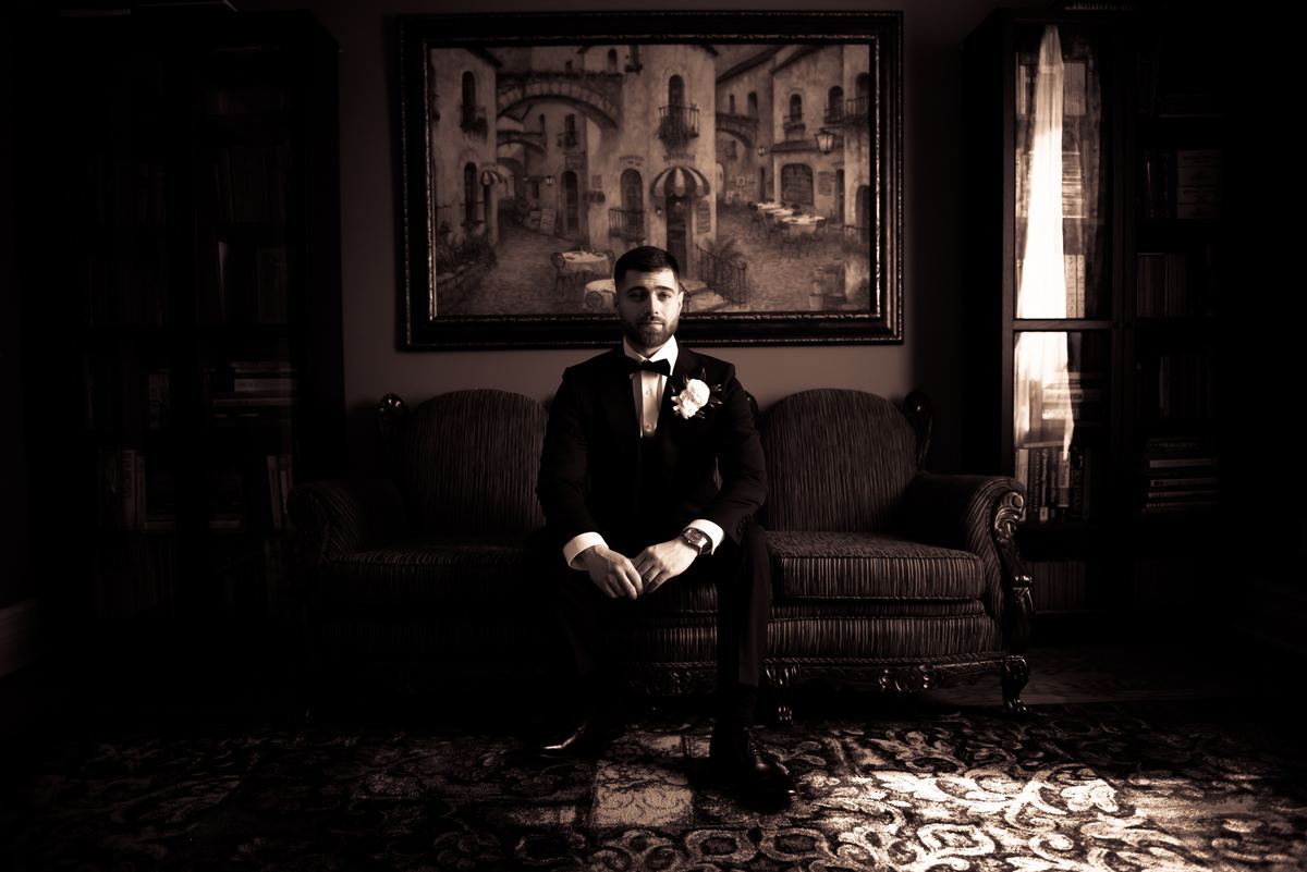 A groom in formal attire sits solemnly on an ornate sofa, illuminated by a single beam highlighting him.