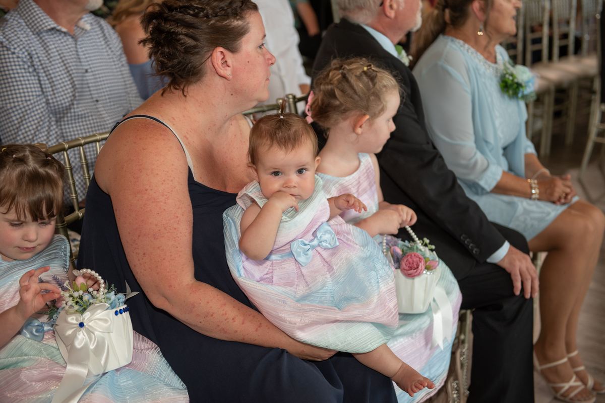 A woman holds a baby in a pastel gown with young girls sitting beside her, each holding floral baskets.