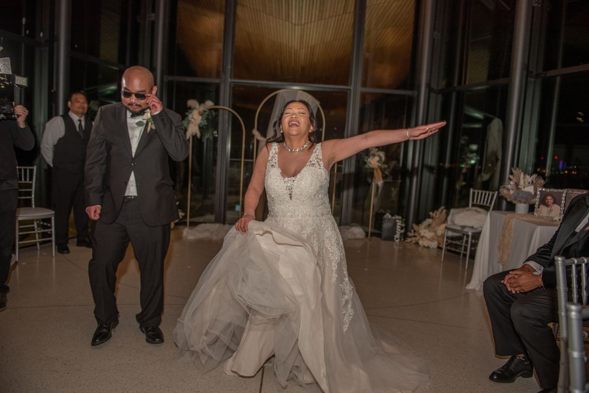 A bride in a tulle-trimmed white dress dances while her groom in sunglasses mirrors her move, tall windows glowing behind.