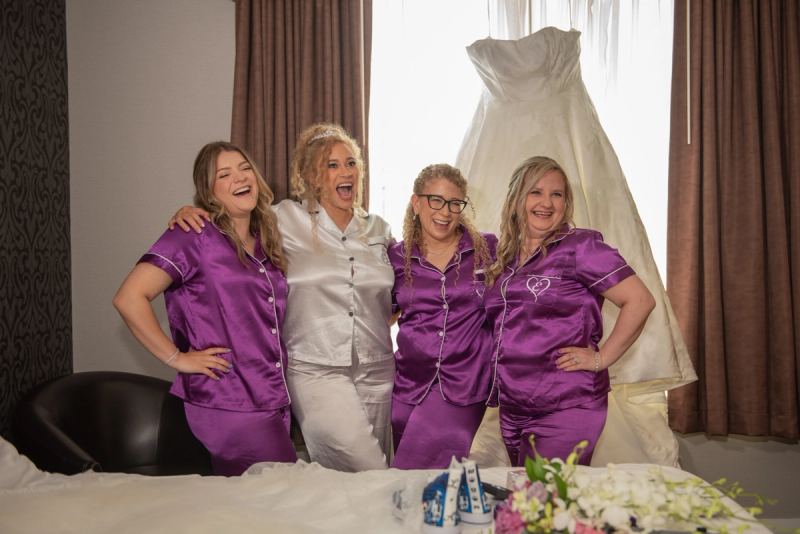 Four women in matching pajamas laugh joyfully near a window with a wedding gown hanging behind them.