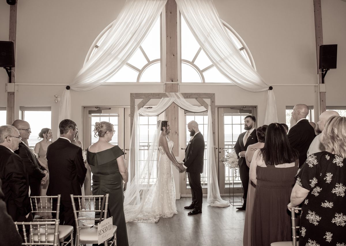 Bride and groom stand at an altar with large arched windows behind them, guests seated in rows, natural light streaming.