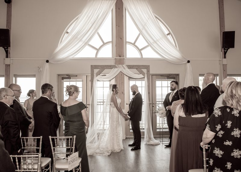 Bride and groom stand at an altar with large arched windows behind them, guests seated in rows, natural light streaming.