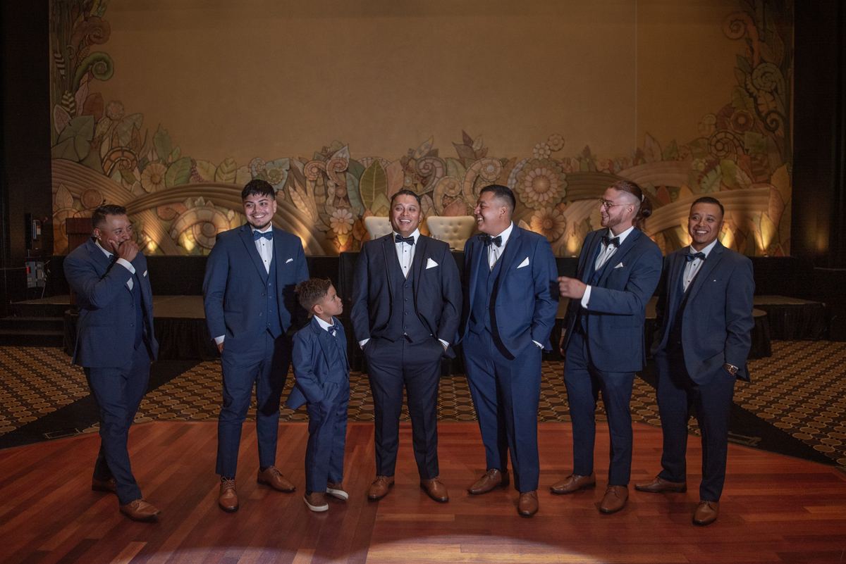 Seven men, including a young boy, laugh in navy tuxedos on a wooden floor with ornate walls during a celebration.
