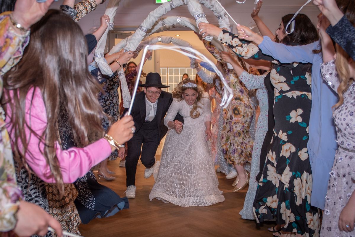 A bride and groom walk through a festive ribbon arch surrounded by smiling guests in colorful attire.