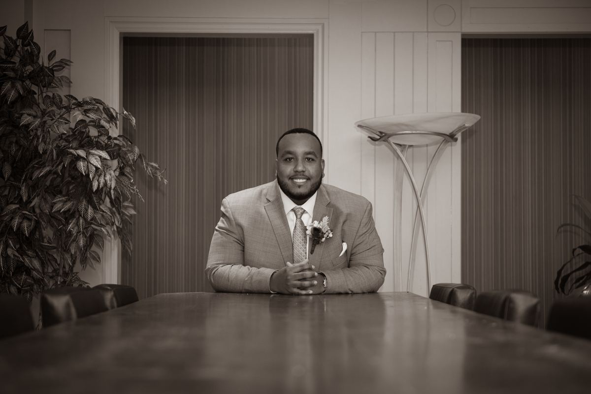 A man in a suit with a boutonnière sits at a long conference table, reflecting in a formal, dimly lit room.