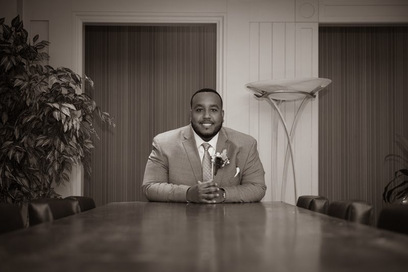 A man in a suit with a boutonnière sits at a long conference table, reflecting in a formal, dimly lit room.