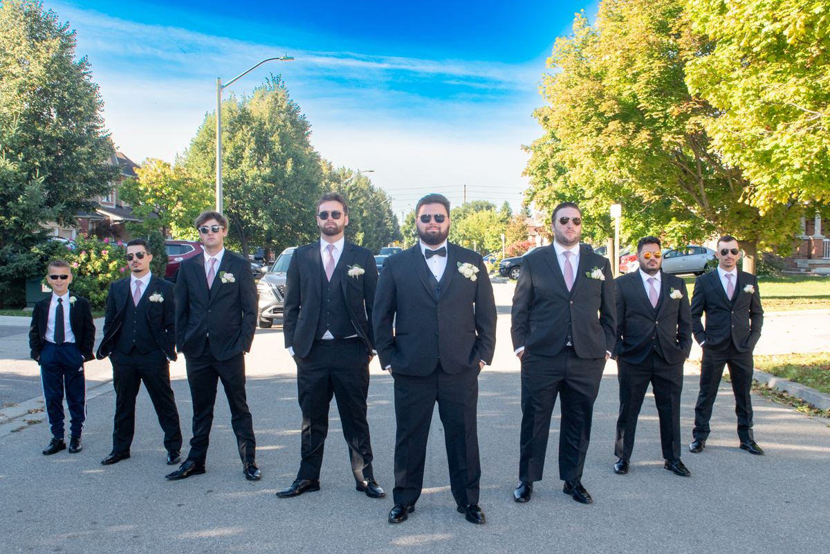 A groom and his groomsmen, including a young boy, pose in formal suits on a suburban street under autumn sunlight.