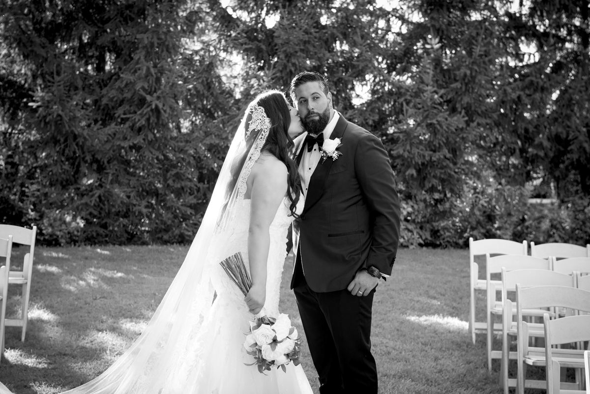 Bride kisses groom's cheek during an outdoor wedding ceremony with sunlight filtering through trees.
