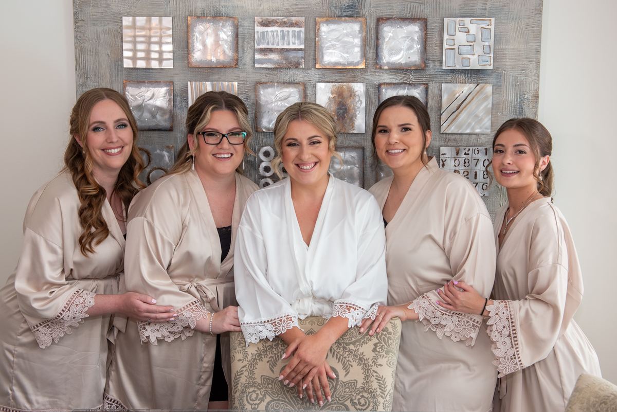 Five women in matching satin robes with lace trim, one in white, smile together before a textured wall art background.