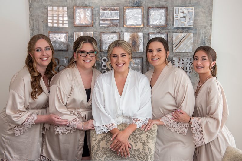 Five women in matching satin robes with lace trim, one in white, smile together before a textured wall art background.