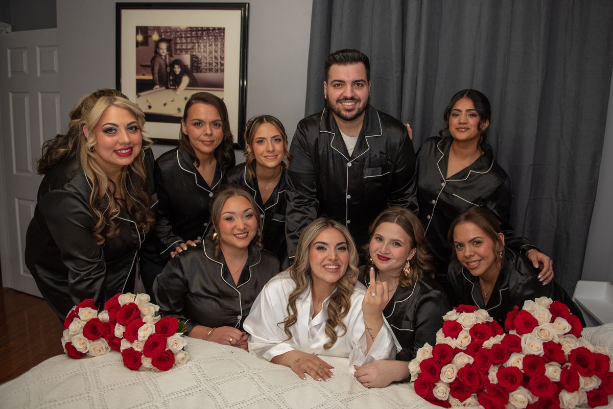 Bride and bridesmaids in matching pajamas gather around bed with rose bouquets during pre-wedding celebration.