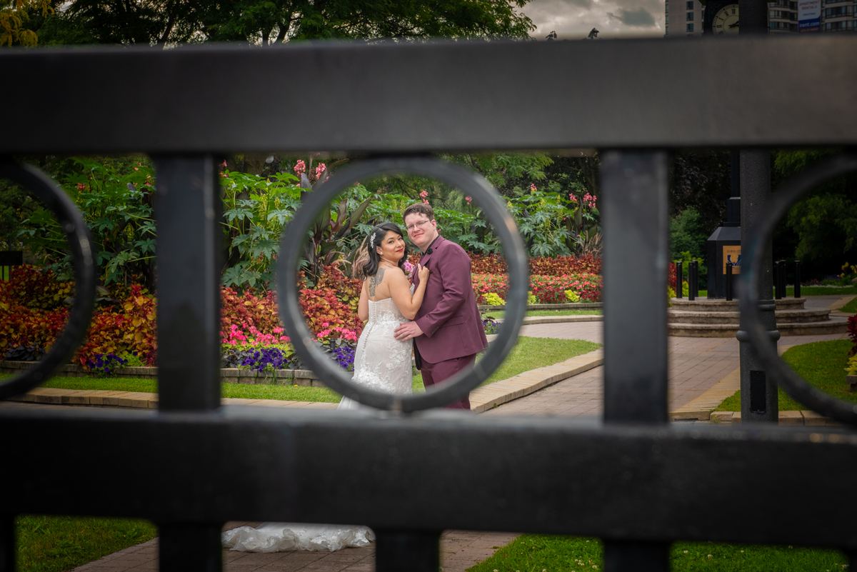 A bride and groom embrace through the circular openings of a dark metal fence in a garden, surrounded by colorful foliage.