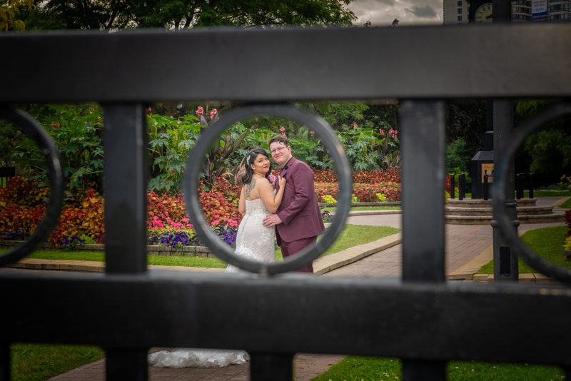 A bride and groom embrace through the circular openings of a dark metal fence in a garden, surrounded by colorful foliage.