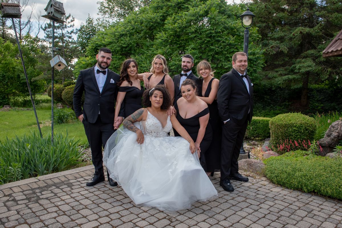 A bride in a white gown sits with her wedding party dressed in black-tie attire amid a lush garden setting.