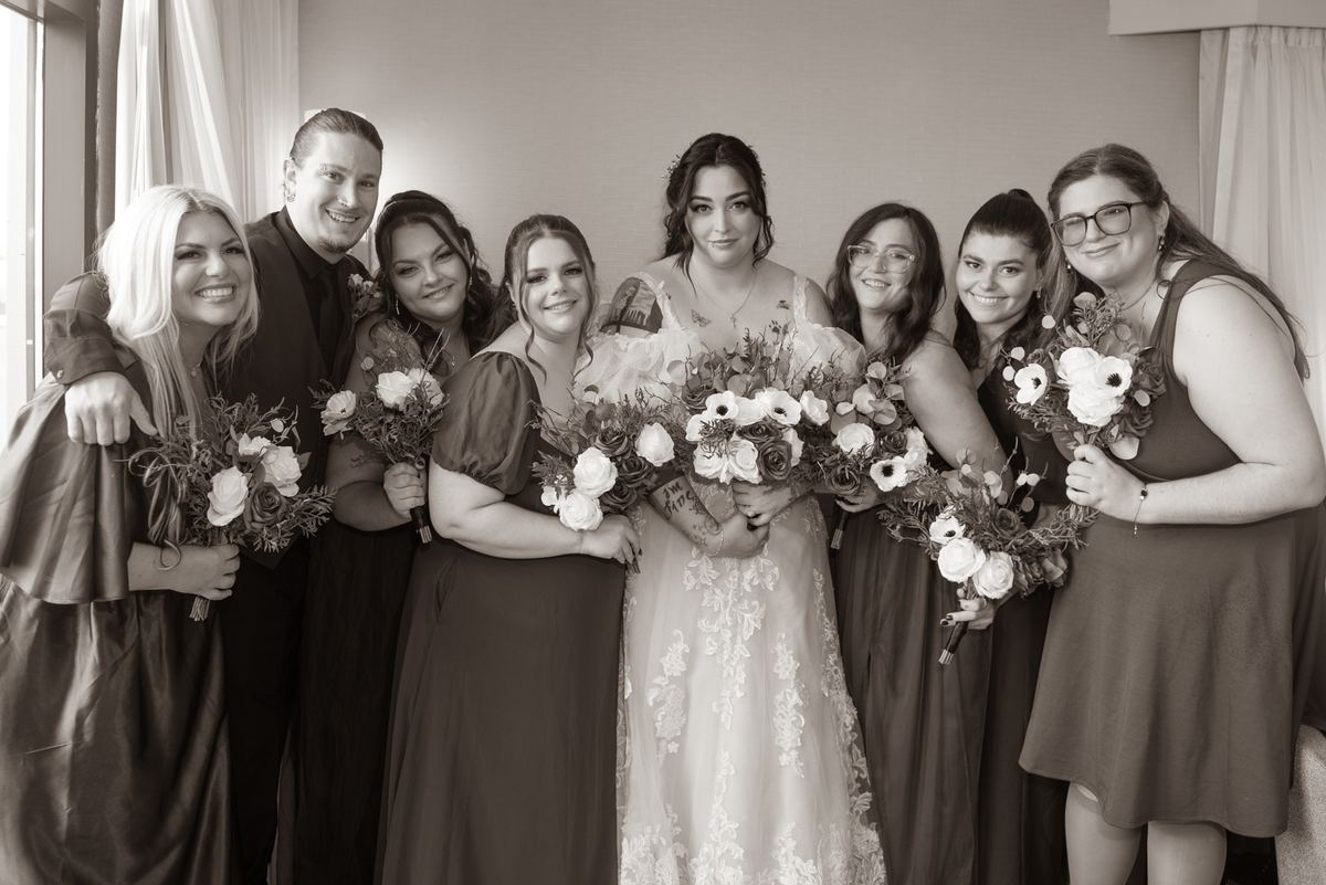 A bridal party gathers closely, smiling and holding bouquets, with soft natural light creating an intimate atmosphere.