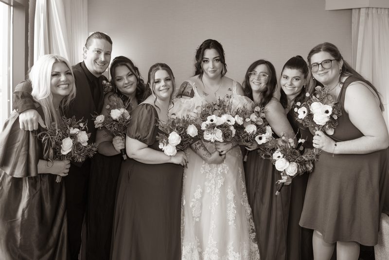 A bridal party gathers closely, smiling and holding bouquets, with soft natural light creating an intimate atmosphere.