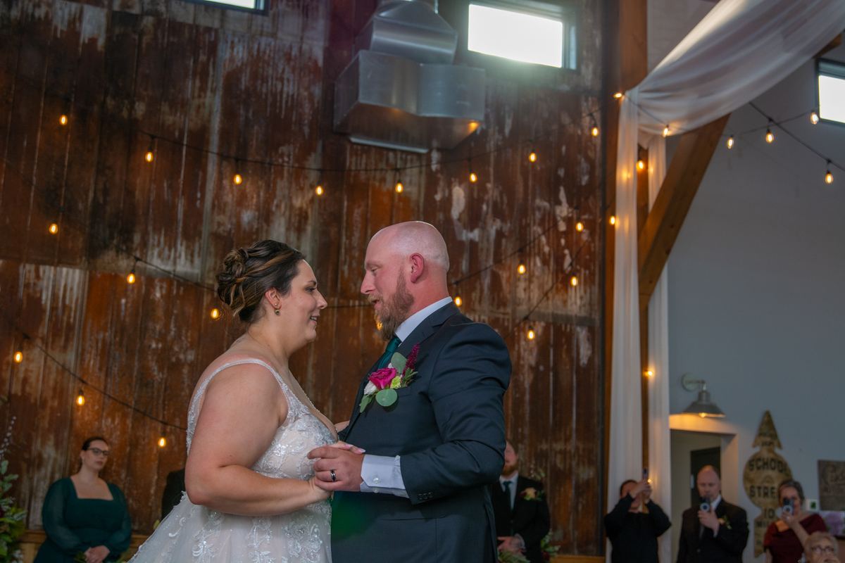 A bride in a white dress and a groom with a boutonniere stand face to face inside a rustic barn hung with string lights.