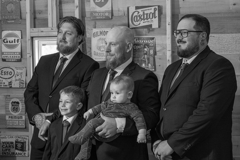 Men and children in suits stand before a rustic wall with vintage car signs during a wedding photoshoot.