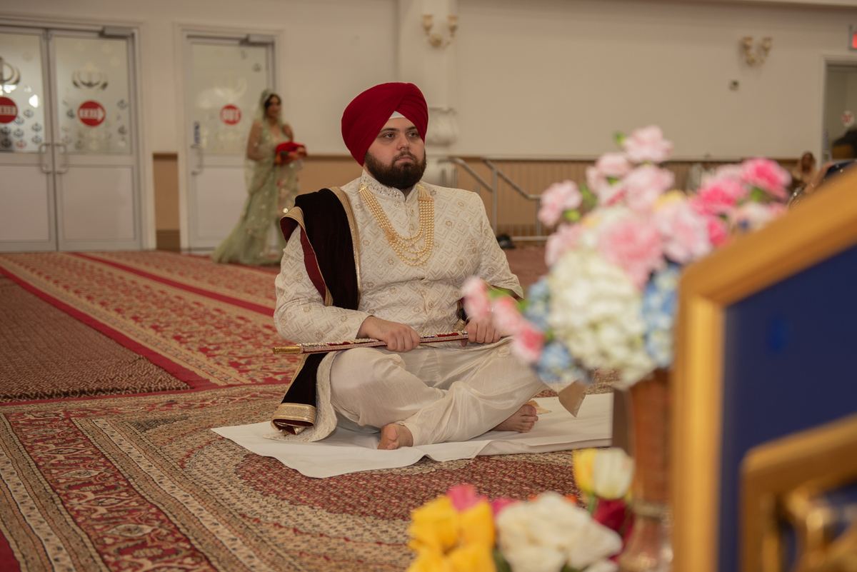 A Sikh groom in white and gold sits cross-legged on a patterned carpet as a bride in green approaches.