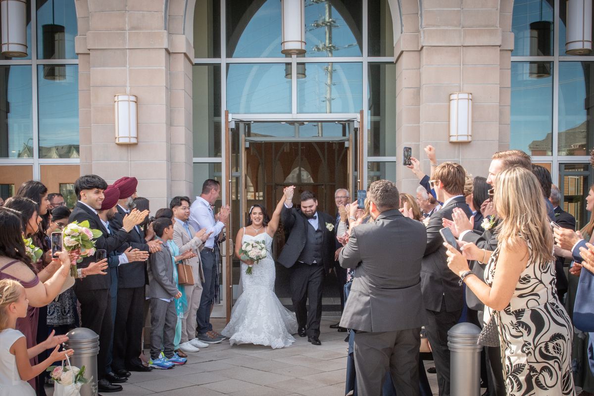 A joyful bride and groom exit a modern stone building with arms raised as guests line the walkway, applauding.