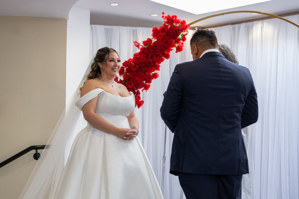 A bride in an off-the-shoulder gown and veil smiles beside a staircase under an arch of red flowers, her groom behind her.