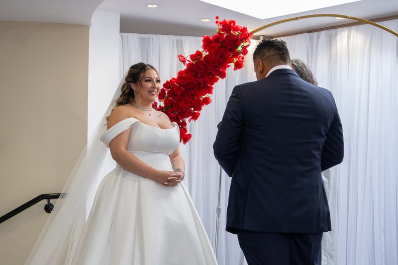 A bride in an off-the-shoulder gown and veil smiles beside a staircase under an arch of red flowers, her groom behind her.
