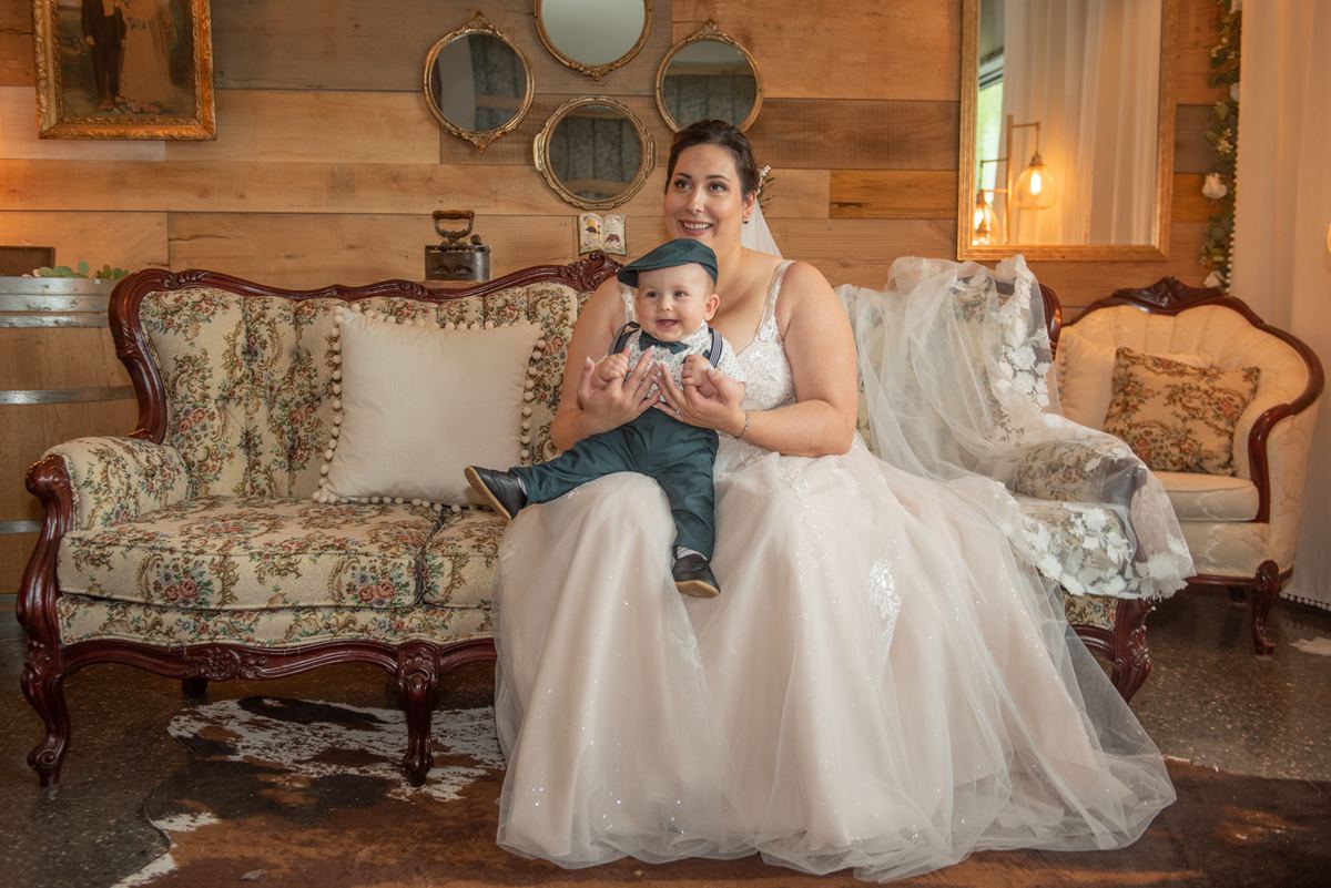 A bride in white holds her baby boy dressed in a tiny suit and bowtie on an ornate sofa in a cozy room.