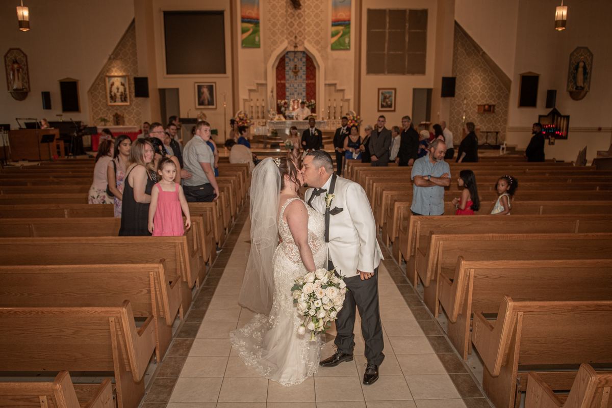 A bride and groom kiss at the altar in a warmly lit church, surrounded by guests and children, with wooden pews.
