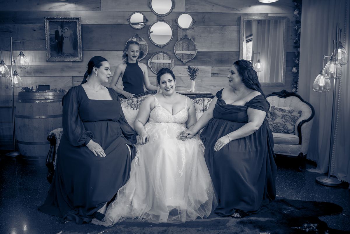 A bride smiles while holding hands with two women in dark gowns, seated at a rustic wooden table under vintage lamps.