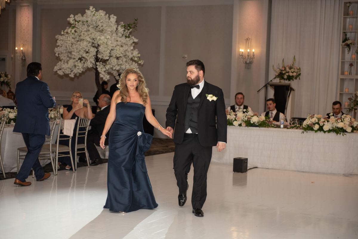 A bride and groom walk across a white dance floor toward the camera during their reception, surrounded by chandeliers.