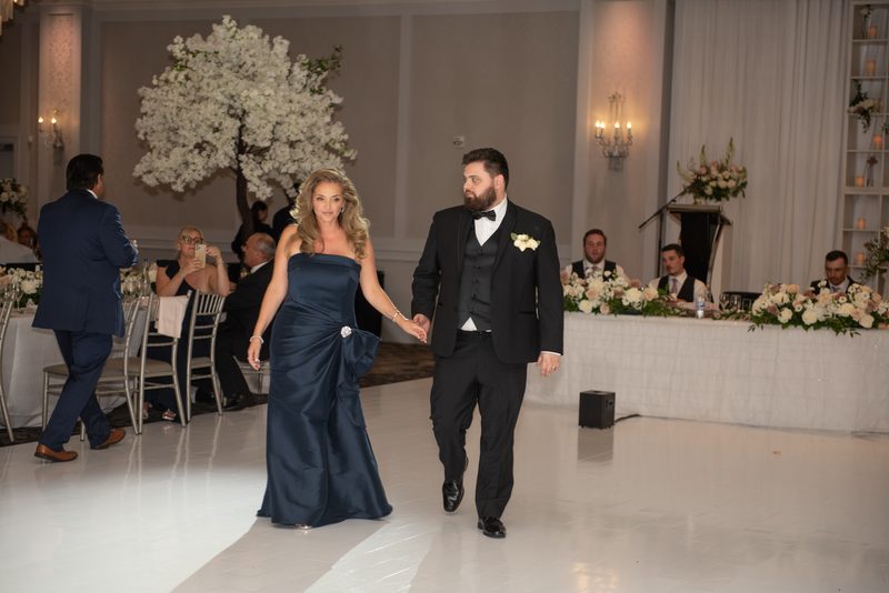 A bride and groom walk across a white dance floor toward the camera during their reception, surrounded by chandeliers.