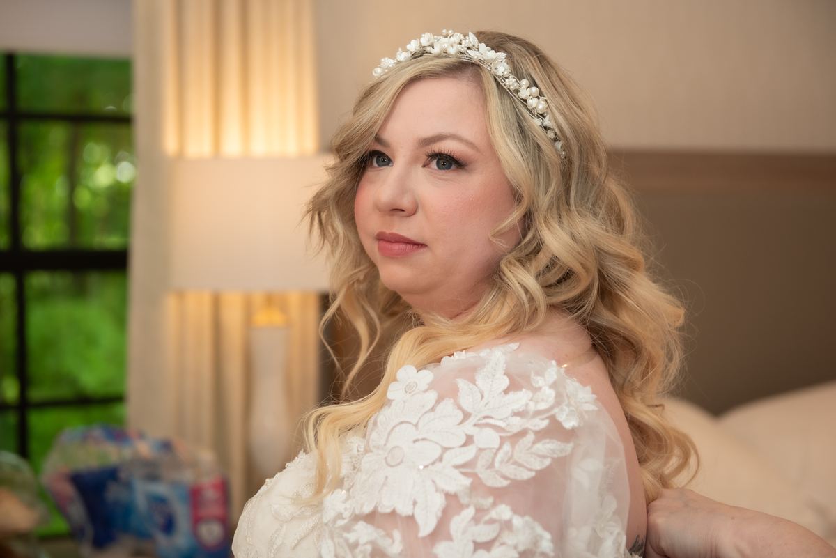 A bride with golden curls and a pearl headpiece looks over her shoulder, illuminated by soft light from a lamp.