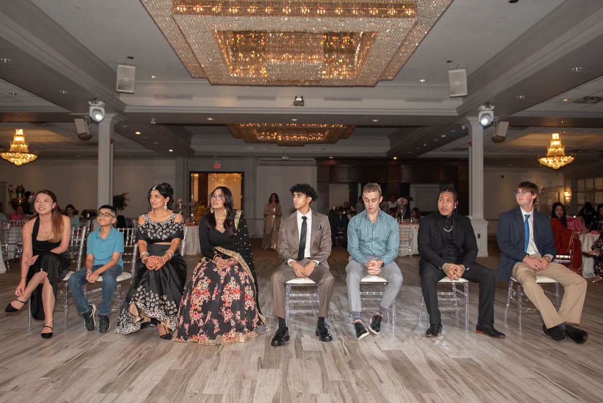 Young adults in formal attire sit on clear acrylic chairs, facing a large crystal chandelier in a grand hall.
