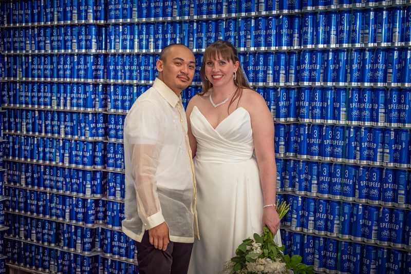 A newlywed couple in traditional and modern attire poses for a portrait against a backdrop of blue beverage cans.