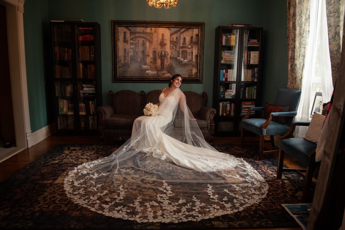 A bride in a white gown and lace veil sits on a vintage sofa in a book-lined room, smiling at the camera.